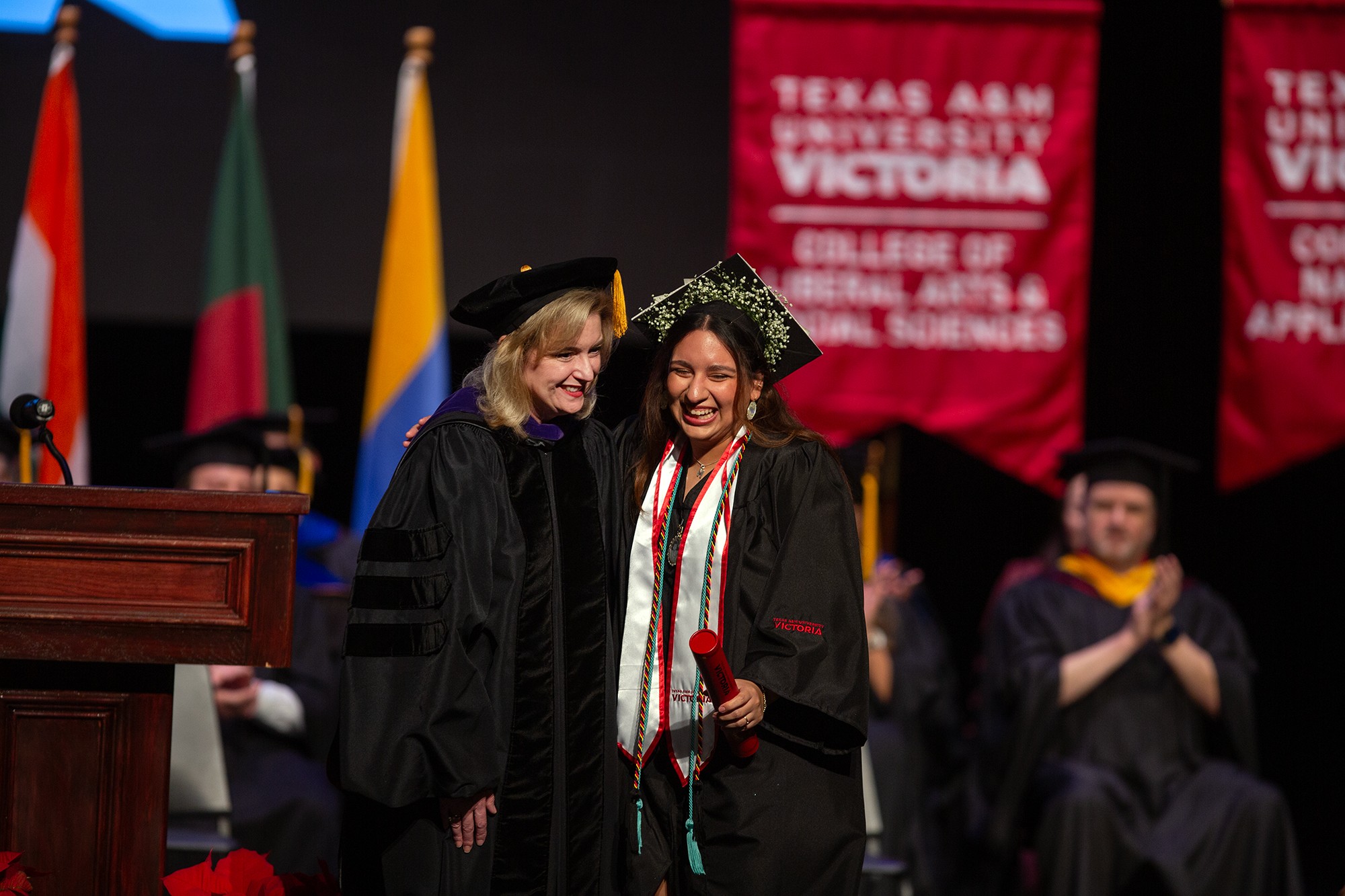 Two women in graduation robes stand on a stage together.